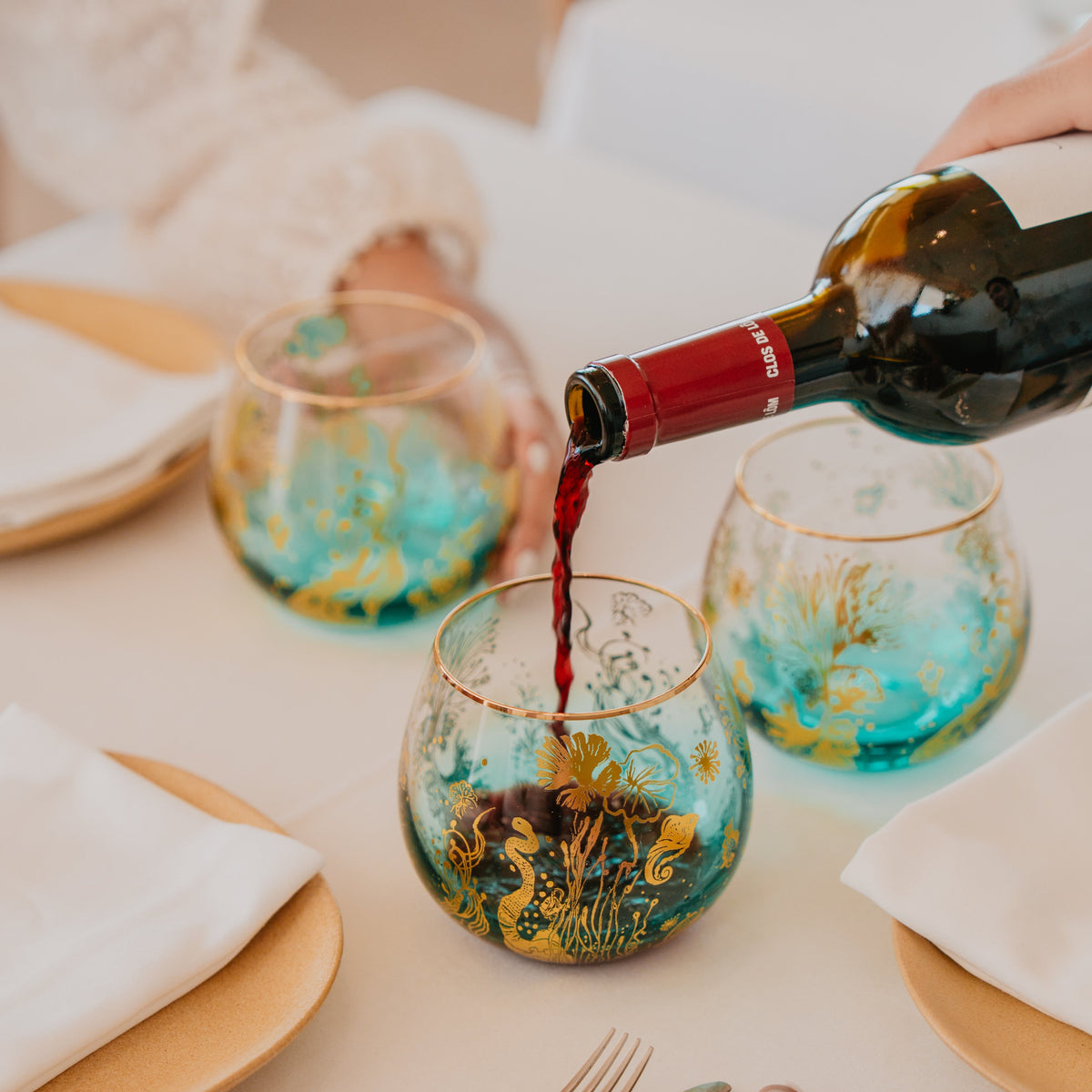 A close-up lifestyle of you pouring red wine from a bottle into elegant stemless wine glasses. 